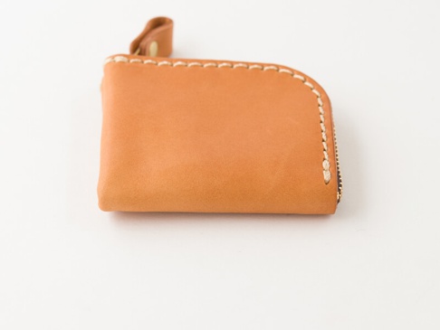 淺棕色皮革製成的手作拉鍊零錢包，邊緣飾有米白色縫線，設有皮革拉環。