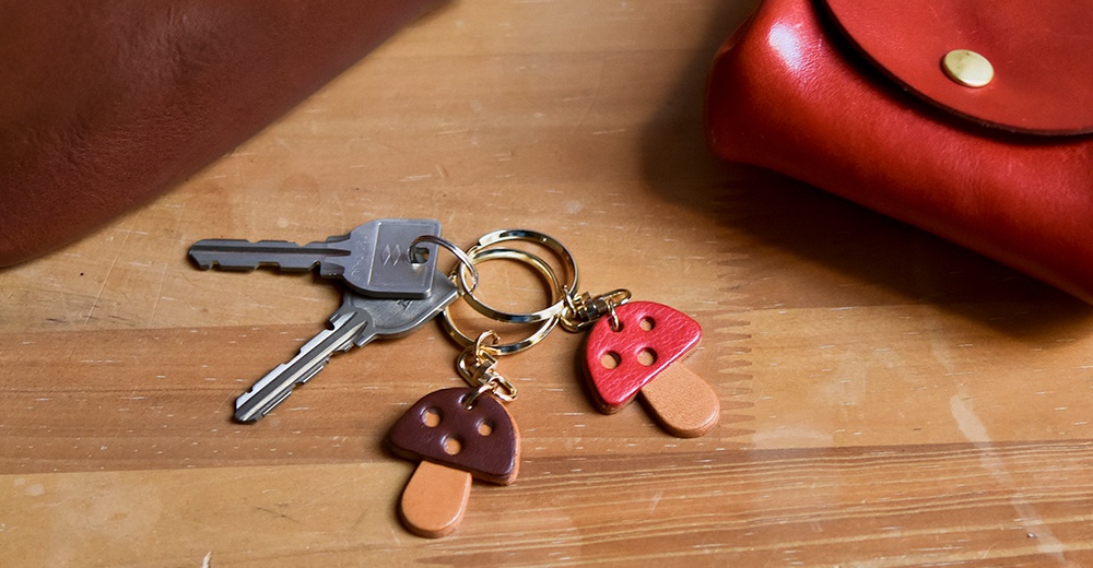 一串鑰匙上掛著兩個職人手作皮革香菇造型吊飾，一個為紅褐色，另一個為深棕色，皆有四個小圓洞，搭配金色鑰匙圈。
