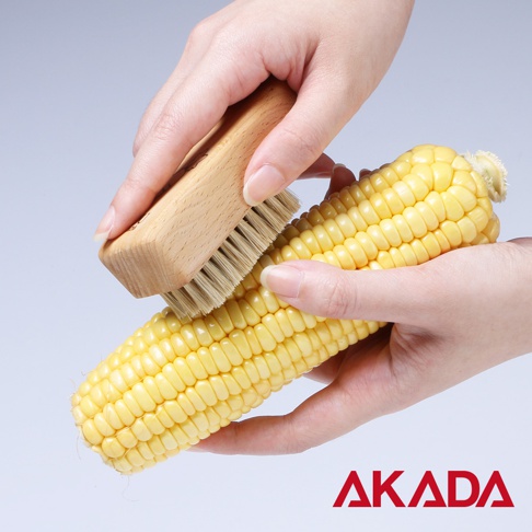 一隻手握著一顆黃色玉米，另一隻手使用木質握柄、豬毛刷毛的蔬果刷輕刷玉米。