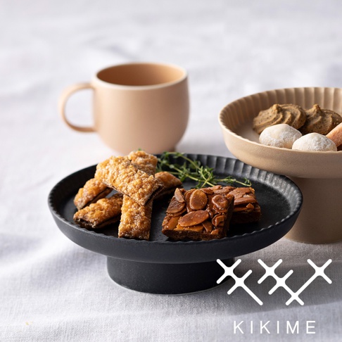 黑色美濃燒高腳平盤，盤上盛有杏仁脆餅、手指餅乾及香草裝飾，背景為淺褐色馬克杯與另一個米白色高腳盤。