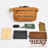 職人手作皮革兩用腰掛斜背包，展示了棕褐色皮革材質、雙拉鍊前袋、可調節腰帶，以及搭配的黑色無線耳機盒、綠色收納袋、橄欖綠鑰匙包、黑色皮夾和棕色長夾。