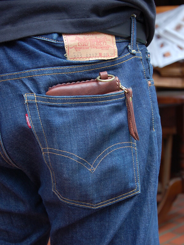 深棕色皮革製成的短夾，帶有獅鬃狀縫線和圓形金屬環，半露出於藍色牛仔褲的後口袋中。