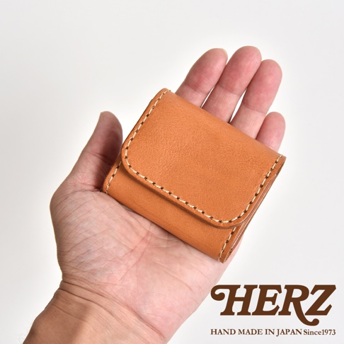 一隻手掌攤開托著一個淺棕色日本HERZ職人手作皮革零錢包，呈現雙折式設計，縫線細緻工整，展現皮革質感與簡約風格。