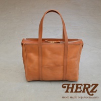 焦糖色皮革製成的肩背拉鍊托特包，包身有縫線細節與一個外層口袋，搭配同色系皮革提把，整體呈現簡約風格。