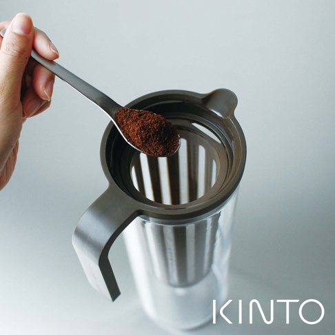 一只手拿著裝有咖啡粉的不鏽鋼湯匙，準備將咖啡粉倒入 PLUG 咖啡冷泡壺的深棕色濾網中。冷泡壺主體為透明玻璃，搭配深棕色霧面把手。