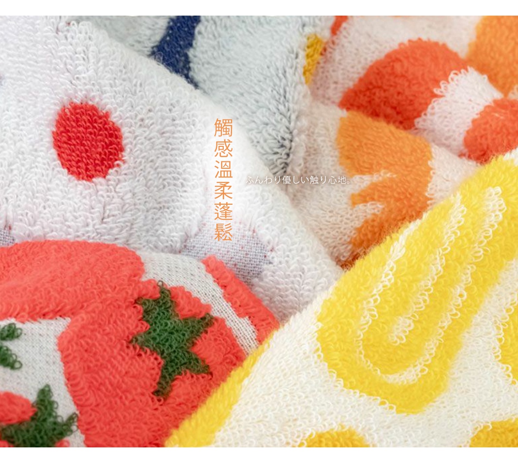 這是一張特寫圖片，展示了幾塊不同顏色的毛巾疊放在一起。毛巾材質柔軟，帶有提花圖案，有紅色的圓點、橙色的花朵和黃色的螺旋形狀，以及綠色的葉子裝飾。其中一塊毛巾上用繁體中文寫著「觸感溫柔蓬鬆」，旁邊還有日文「ふんわり優しい触り心地」。