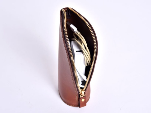 職人手作皮革直立式收納筆袋，呈現自然的棕色皮革質感，拉鍊為金色。袋內裝有白色充電器和淡黃色充電線。