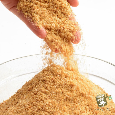 一隻手捧著淺棕色的細小木屑，正從手中傾倒，木屑飄落在白色背景下的玻璃碗中堆積成小山。