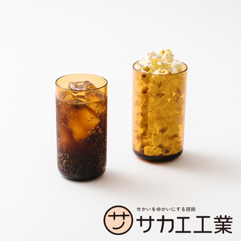 一棕色玻璃杯裝著可樂和冰塊，旁邊另一個棕色玻璃杯裝滿爆米花，下方是SAKAE公司的標誌和日文。