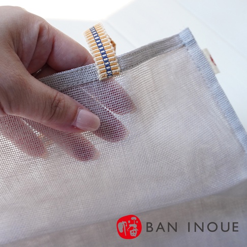 一隻手拿著一個淺灰色網狀收納袋的提把，提把上有藍黃色條紋織帶。收納袋材質為細網布，透氣性佳。