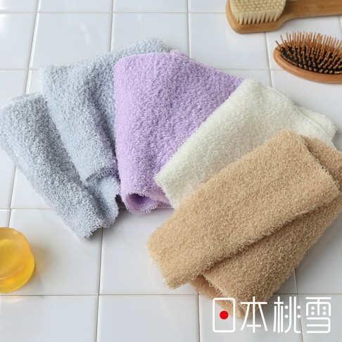 四條不同顏色的沐浴巾，包括淺灰色、淺紫色、米白色和淺棕色，皆為綿柔材質，疊放在白色磁磚背景上，旁邊有一塊透明黃色香皂。