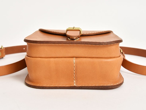 淺棕色皮革翻蓋迷你單肩包，設有金色金屬扣，搭配皮革肩帶，包身線條簡潔，手工縫線清晰可見。