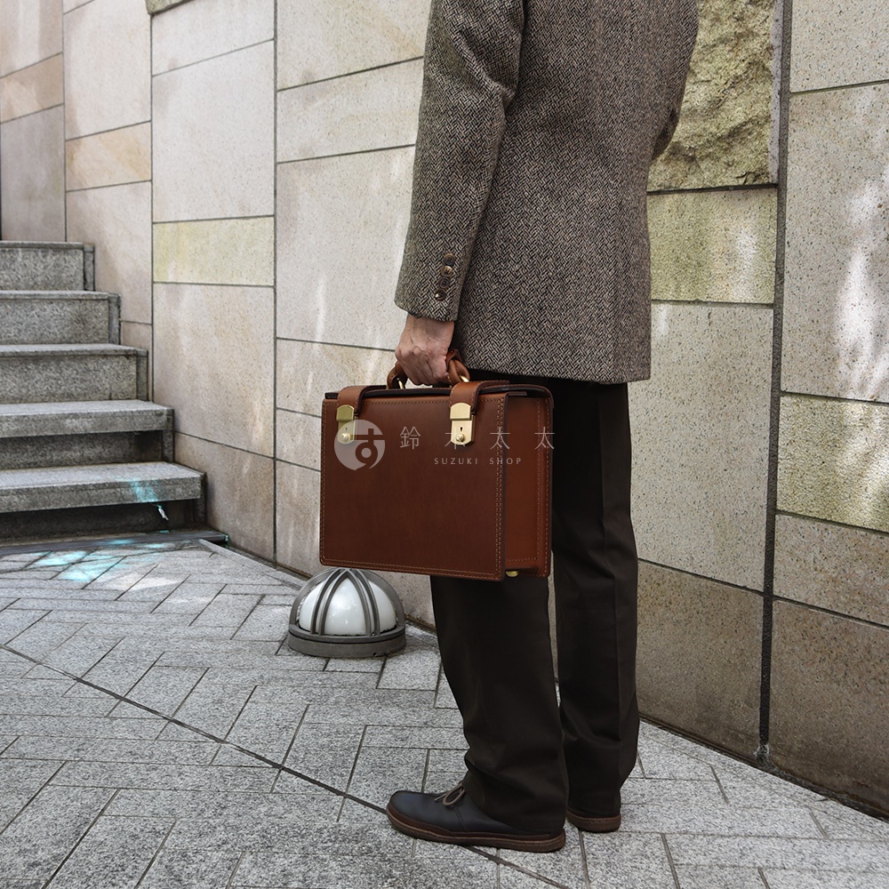 職人手作皮革機師箱，棕色皮革，配有金色金屬鎖扣和提把，側面有縫線細節，適合商務人士使用。