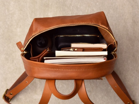 棕褐色皮革後背包，頂部拉鍊敞開，內有書籍、錢包、水瓶和黑色拉鍊零錢包。