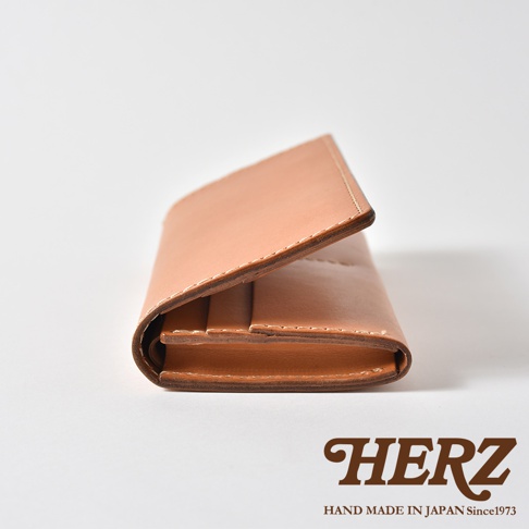 一個採側面視角的日本HERZ職人手作皮革長夾，呈現簡約的棕色皮革質感，翻蓋處與邊緣細節清晰可見，底部印有HERZ HAND MADE IN JAPAN Since1973品牌字樣。