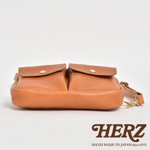 職人手作皮革雙口袋斜背包，淺棕色皮革，正面有兩個附金色釦的翻蓋口袋，側邊有可調節的皮革背帶和金色金屬扣環。