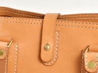 淺棕色皮革托特包局部特寫，展示精緻縫線、皮革翻蓋及黃銅色原子釦，材質為天然皮革。