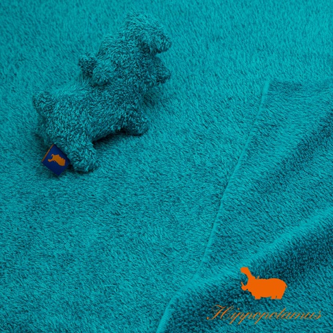 藍綠色竹棉混紡布料上，有一個藍色標籤的淺藍色河馬造型抓握玩偶，玩偶材質柔軟，布料表面有絨毛感，右下角有橘色河馬圖案及品牌Logo。