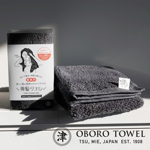 深灰色毛巾，觸感蓬鬆，質地柔軟，疊放在白色表面上，旁邊是產品包裝盒，上面印有手繪圖案和日文說明。