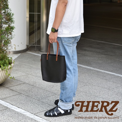 一位身著白色上衣與淺藍色牛仔褲的男子，手提一個由日本HERZ品牌職人手作的黑色皮革直式托特包，包款配有對比鮮明的棕色手把，男子腳穿黑色涼鞋，畫面右下角帶有HERZ HAND MADE IN JAPAN Since1973的字樣。