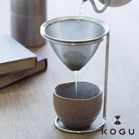 日製18-8不鏽鋼濾茶器，圓錐形細網濾網，搭配銀色金屬立架，正將茶湯濾入下方陶製茶杯中，背景有書本與金屬壺。