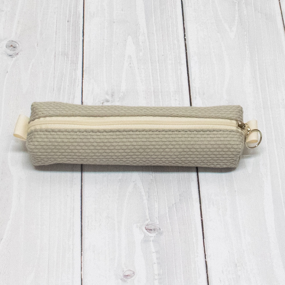 米色三河木綿刺子織筆袋，帶有米色拉鍊和金色拉環，放置於淺色木紋背景上。