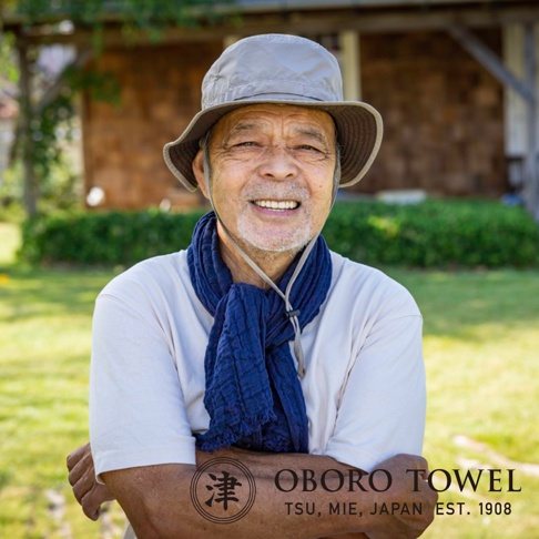 一位年長男性戴著灰色漁夫帽，身穿白色T恤，頸部圍著一條深藍色三層紗純棉輕柔掛脖毛巾，背景是戶外綠草地與建築。