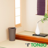 灰色圓形半開式垃圾桶，底部較寬，向上逐漸收窄，頂部設有半圓形開口，垃圾桶放置於日式榻榻米地板上，背景為淺色牆壁及木框窗戶。