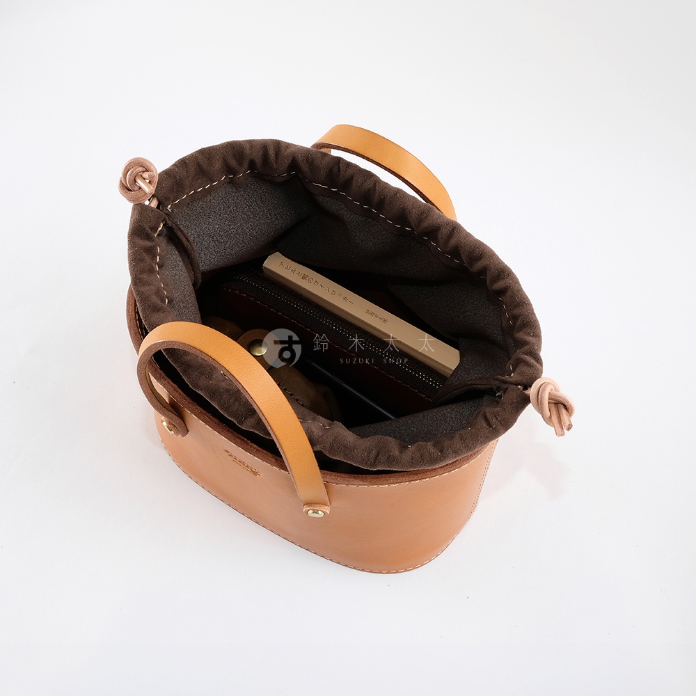 淺棕色皮革束口手提包，內裡為深棕色帆布，配有兩個短提把，包口處有抽繩設計，包內可見一個皮夾、一支筆及一本書。