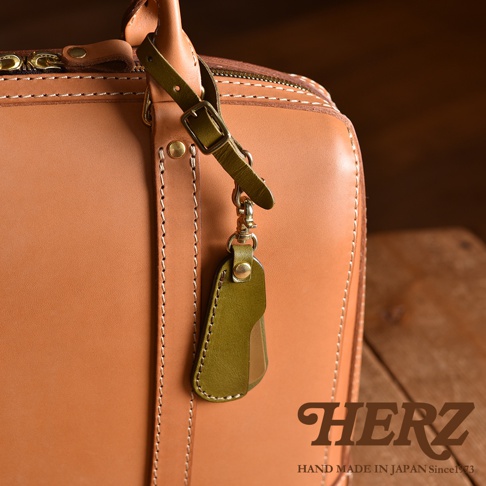 職人手作皮革隨身金屬鞋拔，附有橄欖綠皮革掛飾，搭配淺棕色皮革箱包。