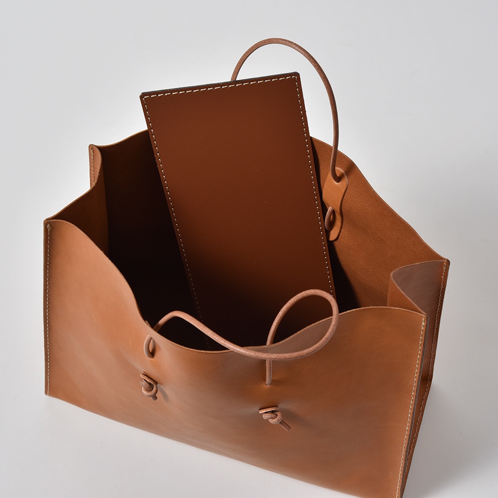 職人手作皮革提袋 T-5，採用鞣製皮革製成，顏色為溫潤的棕色。提袋為敞口設計，內部附有隔層，兩側有皮革繫帶收納設計，整體線條簡約。