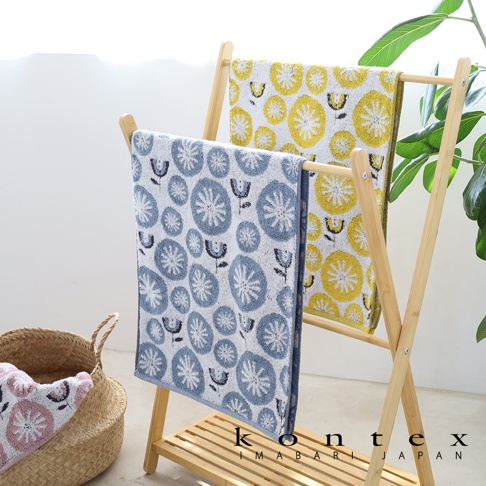在木製毛巾架上掛著兩條印有花朵圖案的毛巾，一條為藍色，一條為黃色，旁邊有一個編織籃裝著另一條粉色花朵圖案的毛巾，底部有KONTEX IMABARI JAPAN的標誌。