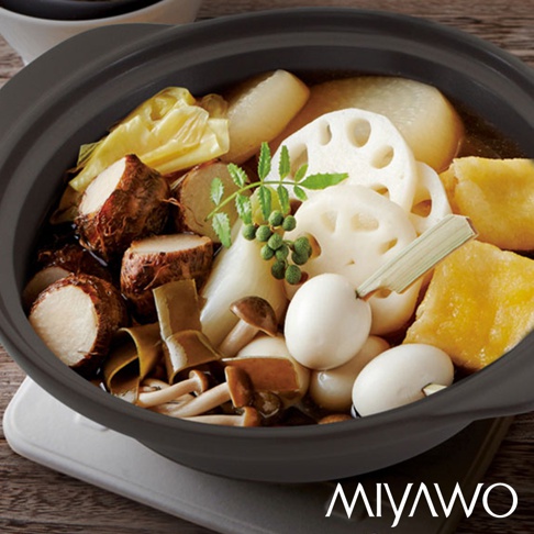 深灰色陶土湯鍋中盛有豐富的日式火鍋料，包括白蘿蔔、芋頭、蓮藕、菇類、鵪鶉蛋及綠色香料，湯汁呈金黃色。