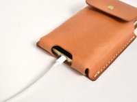 淺棕色植鞣皮革手機收納包，包側縫線明顯，包蓋處有銅質按扣，手機充電線插入其中。