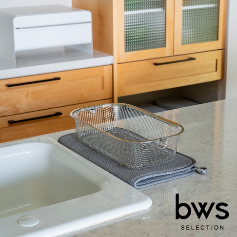 廚房檯面上，一個灰色的可折疊收納瀝水墊上放置著一個銀色的金屬網籃。背景有木質櫥櫃和一個白色的廚房用品。