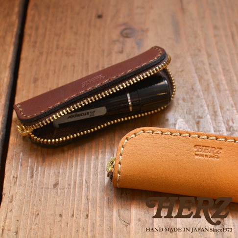 深棕色皮革印章收納袋，拉鍊開合，內部可見印章。右側為淺棕色皮革印章收納袋，同樣為拉鍊設計，袋身壓有品牌標誌。兩款均為手工縫製，材質為皮革。
