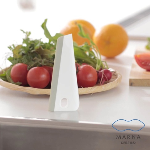 白色廚房清潔刮板，傾斜放置於檯面上，前方有圓形掛孔。背景為一籃新鮮蔬菜，包含番茄、蘿蔔和綠葉菜，以及兩個橘子。