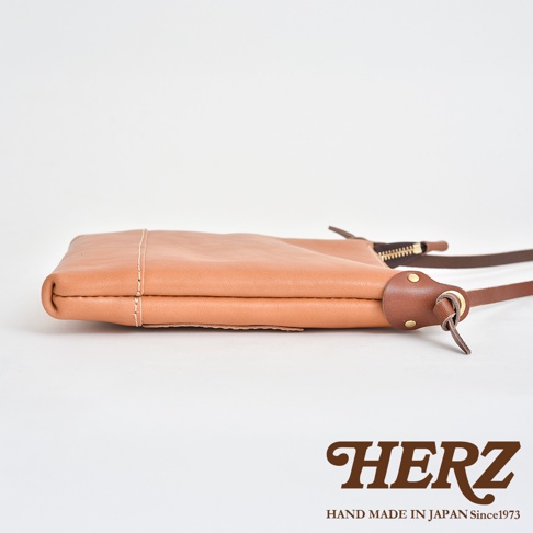 職人手作皮革直式隨身單肩包，淺棕色，側邊展示拉鍊與皮革背帶連接處，材質為真皮。
