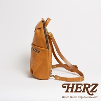 職人手作皮革大開口圓角後背包，呈現自然棕色皮革質感，設有拉鍊主袋及前置拉鍊口袋，搭配可調節的皮質肩帶和金色五金配件。