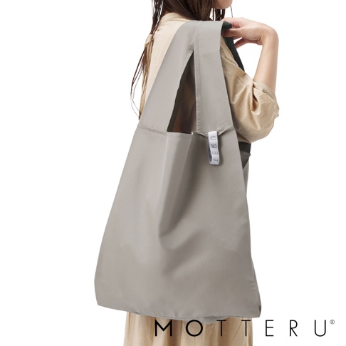 一個女性穿著米色上衣，肩上挎著一個灰色的大容量摺疊環保隨行袋，袋子由尼龍材質製成，側面有一個白色小標籤，上面印有黑色字母「LEKU」。