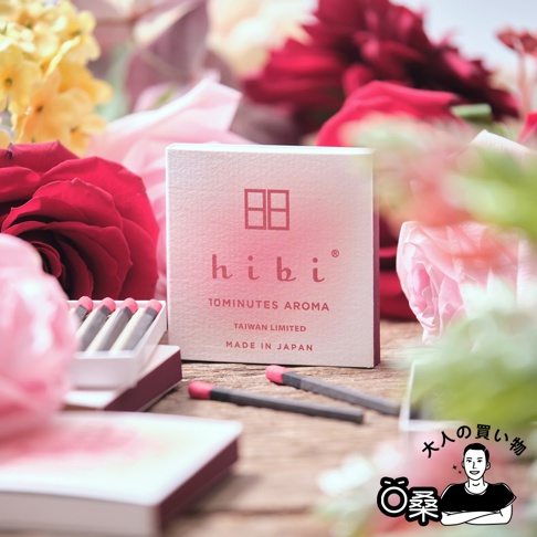粉紅色紙盒裝的香氛火柴，盒上印有「hibi 10MINUTES AROMA TAIWAN LIMITED MADE IN JAPAN」字樣。盒旁散落幾支黑色火柴，火柴頭為粉紅色。背景為粉色、紅色、黃色的花朵。