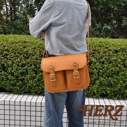 日本HERZ職人手作皮革經典雙扣郵差包，在綠色植被背景下，模特身穿灰色襯衫和牛仔褲，斜挎著一個棕色皮革郵差包，包包上有兩個金屬扣環和兩個前袋，包包底部有HERZ品牌標誌和MADE IN JAPAN Since1973字樣。