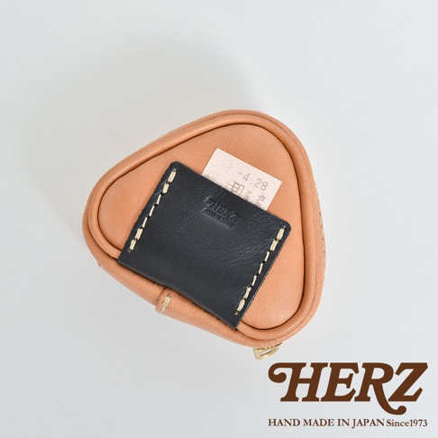 職人手作皮革小物收納包，呈三角飯糰造型，主體為淺棕色皮革，上方覆蓋黑色皮革小片，以米白色縫線裝飾。