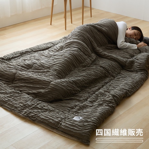 一位年輕女性正側躺在木質地板上，蓋著一條呈現自然皺褶紋理、帶有深灰色調的四國纖維柔滑單人被，畫面展現出舒適溫馨的居家休息氛圍，右下角印有四國纖維販賣的文字標示。