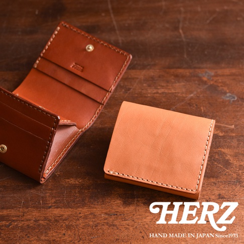 日本HERZ職人手作皮革雙折迷你短夾，展示一個已打開呈現內部夾層結構的皮夾，以及另一個合上的淺棕色皮夾，皆置於深色木紋桌面，右下角印有HERZ品牌標誌與創立年份。