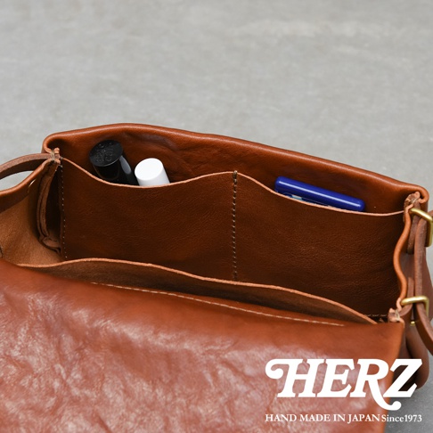 職人手作皮革斜背包，棕色，內部有隔層，可見一支黑色筆、一支白色護唇膏和一支藍色筆。