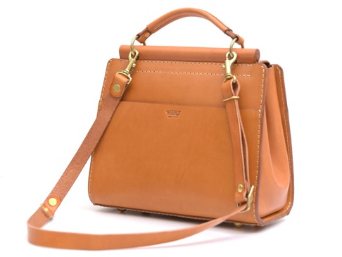 一個棕褐色皮革製成的兩用單肩包，帶有頂部提把、可拆卸肩帶和黃銅色五金配件，包身縫線清晰可見，正面有品牌壓紋。