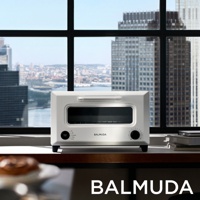 白色BALMUDA烤麵包機，設有玻璃門和銀色旋鈕，置於窗前木桌上，背景為城市高樓景觀。