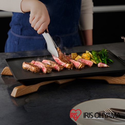 黑色 IHU-A10-B 輕薄電烤盤上，用夾子夾著一塊煎至金黃的牛排，旁邊擺放著香煎馬鈴薯塊、烤蔬菜和蒜頭。烤盤架為木質，整體呈現烹飪美食的畫面。