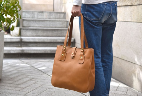 手提式焦糖色皮革托特包，配有兩條皮革提把、前袋和中央按鈕封口。